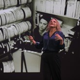 Brainwashed:Sex-Camera-Power. A woman standing on stage, in front of a black and white background, seemingly speaking to a crowd who are not in picture. 