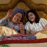 A film still from 'Are you there God? It's me Margaret'. A Grandma and Granddaughter lay in bed laughing at something. 