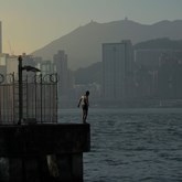Blue Island - a man stands on a pier looking down into the water, against the background of the Hong Kong skyline
