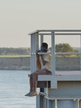 A man is sitting on a pier, speaking on his phone. The Dam film still. 