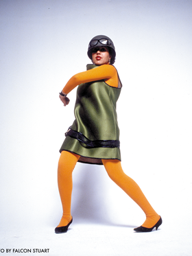 Poly Styrene: I Am A Cliché - woman wearing yellow tights, short green dress and hat stands against white background.