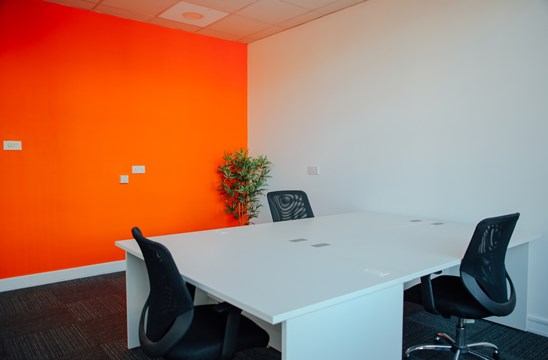 Office 308 with a white desk surrounded by three office chairs and a plant. In the background is a bright orange wall.